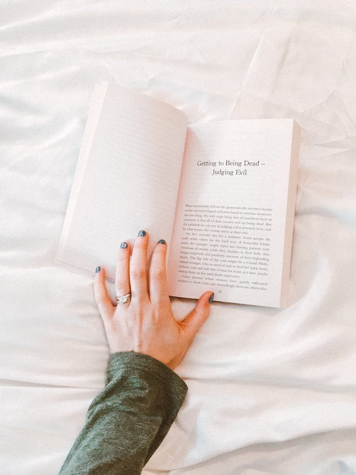 Close-up of a woman's hand holding a book while relaxing on a bed with white sheets. Perfect for lifestyle themes.